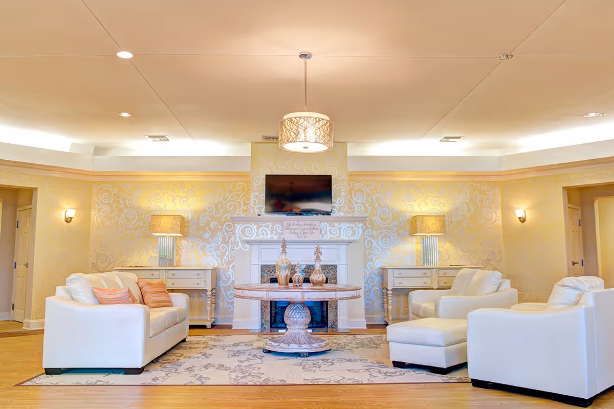 A bright and elegant living room with white leather sofas and armchairs arranged around a round wooden coffee table. The room features a decorative fireplace with a flat-screen TV mounted above it, two matching lamps on side tables, and patterned wallpaper with a light yellow and white swirl design. The floor is wooden with a large area rug under the furniture.