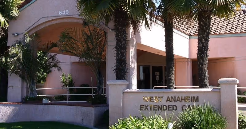 Exterior view of a building entrance with a covered archway, palm trees, and a sign that reads 'West Anaheim Extended Care'. The building has a tiled roof and light-colored walls with some greenery in front.