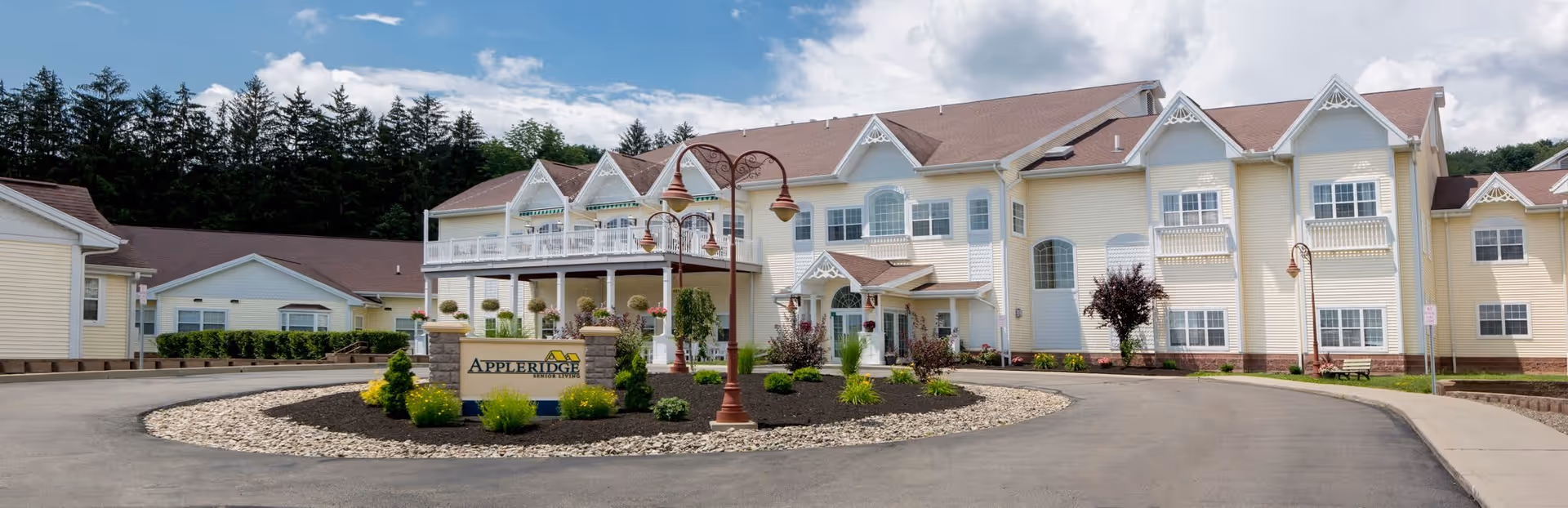Front exterior view of Appleridge Senior Living facility featuring a large, light yellow building with white trim, multiple windows, and a covered entrance. There is a circular driveway with a landscaped island in the center that includes a sign reading 'Appleridge' and decorative street lamps. Trees and a partly cloudy sky are visible in the background.