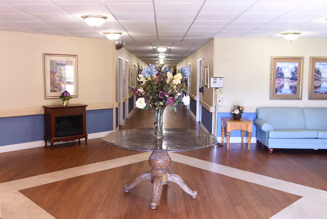 Interior lobby/hallway with a round table holding a vase of flowers centered in front of a long corridor, seating and framed artwork along the walls.