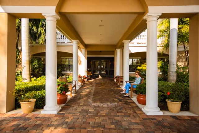 Covered walkway with white columns and brick flooring leading to a building entrance, surrounded by potted plants and greenery. A woman is sitting on a bench on the right side under the covered area.