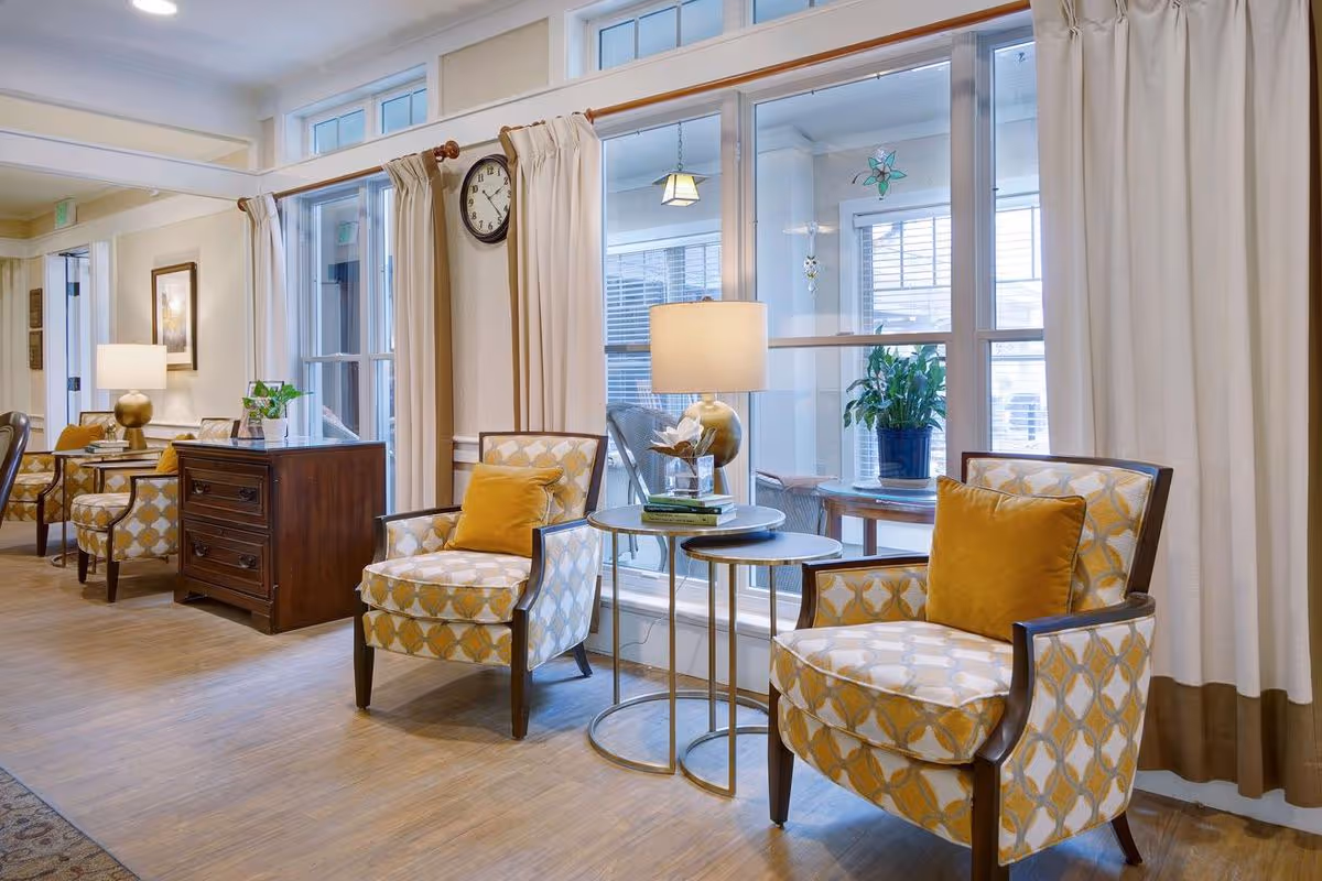 A bright and cozy sitting area in a senior living facility with two patterned armchairs featuring yellow cushions, a small round side table with a lamp and books, large windows with beige curtains, a wooden chest of drawers, and a wall clock.