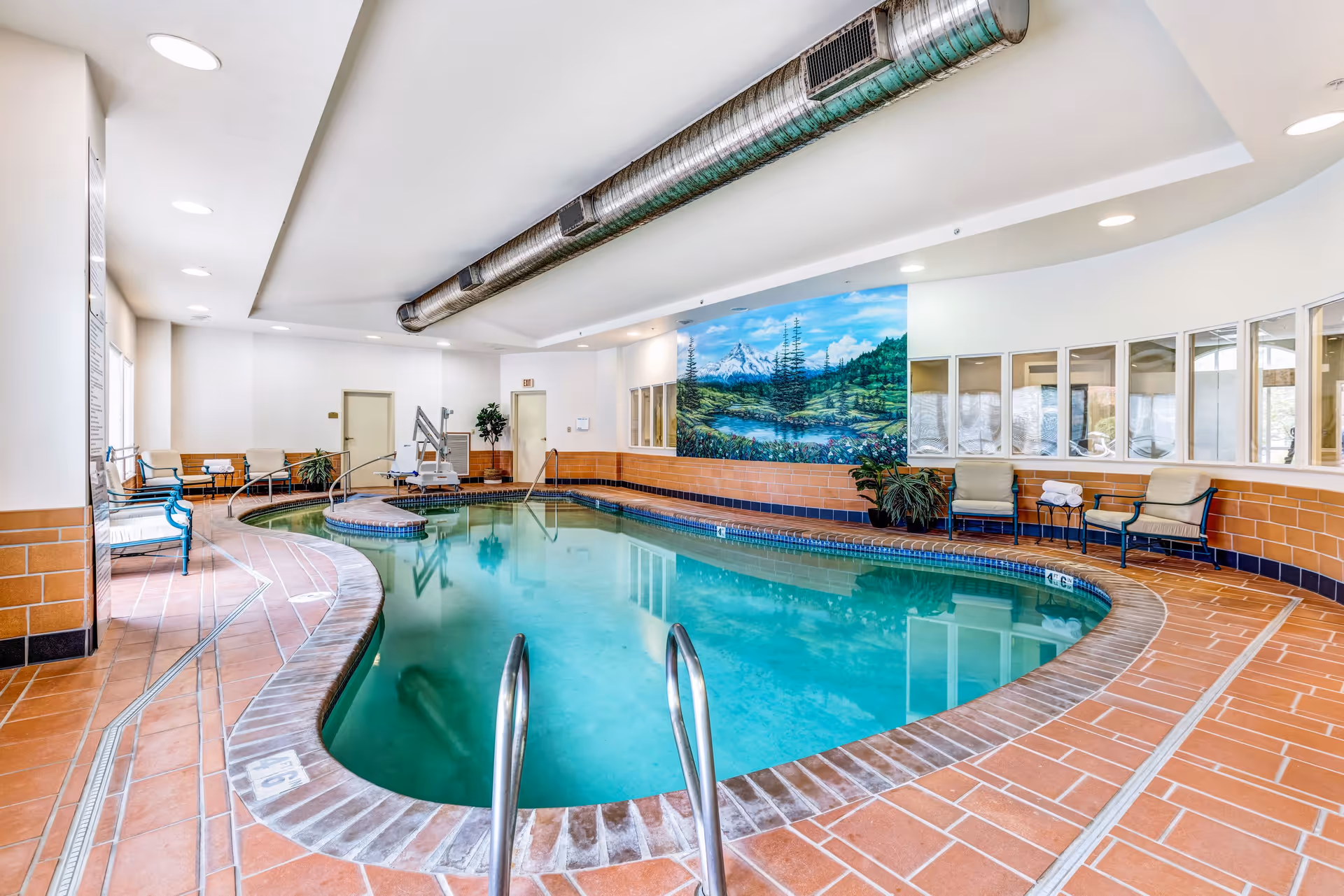 Indoor swimming pool area with terracotta tile flooring and curved edges. The pool is surrounded by chairs and plants, with a mural of a mountain landscape on the far wall. Large windows and ceiling lights illuminate the space.