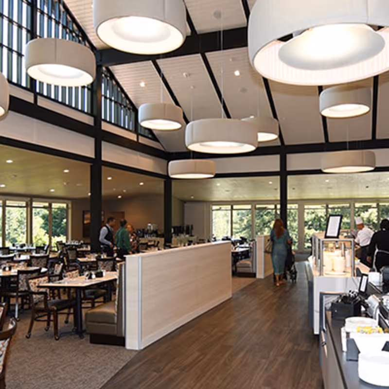 Interior view of a spacious dining room with large windows allowing natural light, modern round pendant lights hanging from a high ceiling, several tables and chairs arranged for dining, and a few people walking and interacting in the background near a food service area.