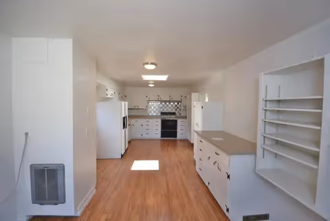 Long bright kitchen with white cabinets, wood floors, and appliances in an open layout.