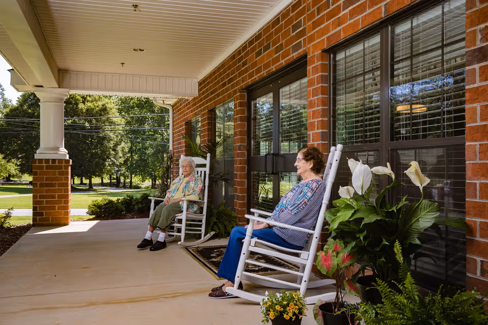 Two elderly women sitting on white rocking chairs on a covered porch outside a brick building. There are potted plants nearby and green trees in the background under a clear sky.