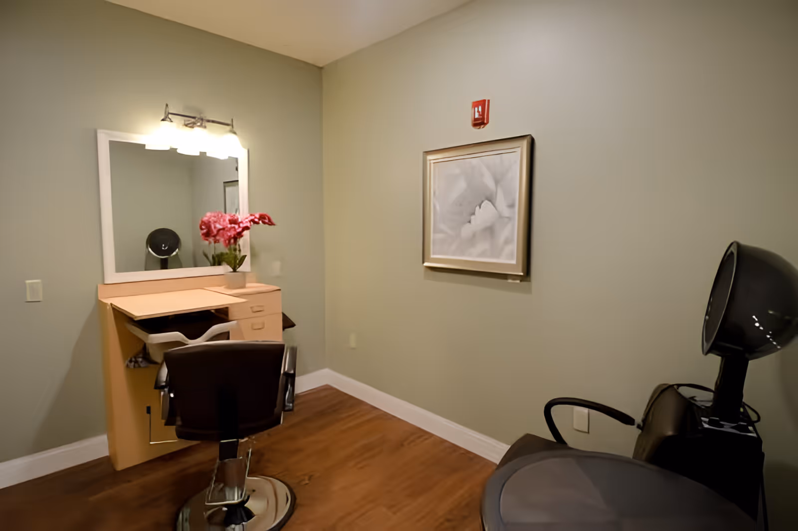 A small salon area with a styling chair in front of a vanity mirror with lights. A vase with pink flowers is placed on the vanity. To the right, there is a black hair dryer chair. The walls are painted light green and there is a framed picture on the wall.