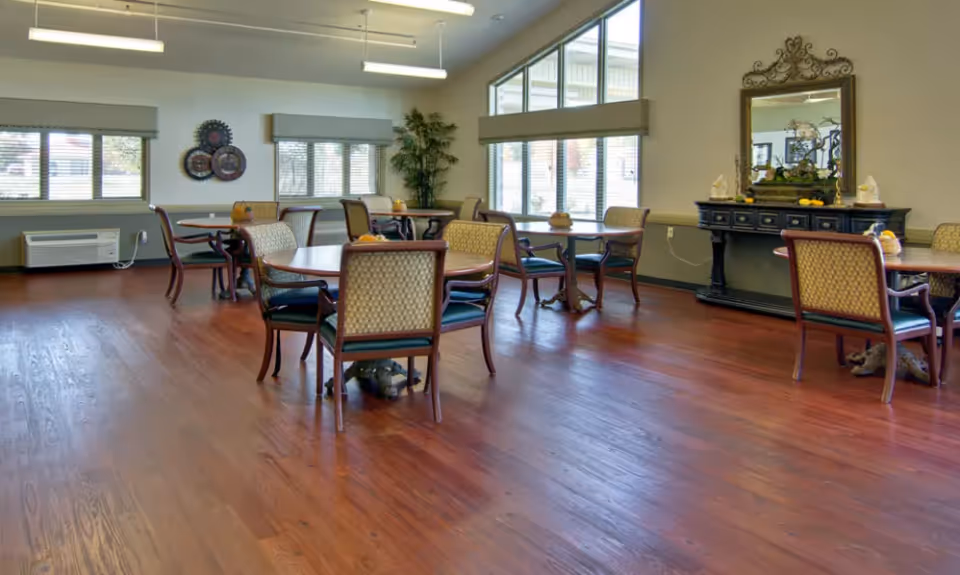 A spacious communal dining room with round tables and chairs on polished wood floors and large windows.