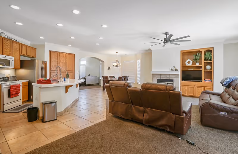 Open-concept living area with a kitchen island, dining table, and brown leather recliners facing a fireplace and built-in shelving.