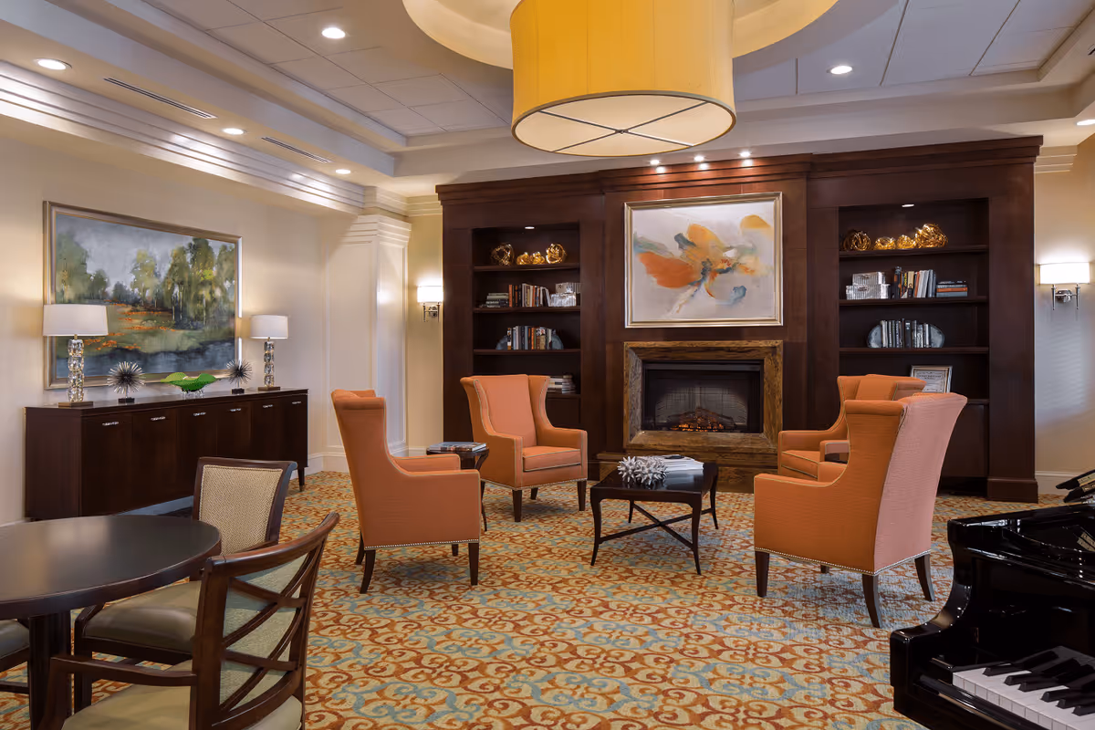 A cozy living room area with four orange upholstered armchairs arranged around a small dark wood coffee table. Behind the chairs is a dark wood built-in bookshelf with decorative items and books, and a fireplace with a colorful abstract painting above it. To the left, there is a sideboard with two table lamps and a landscape painting on the wall. The room has a patterned carpet and a large ceiling light fixture.