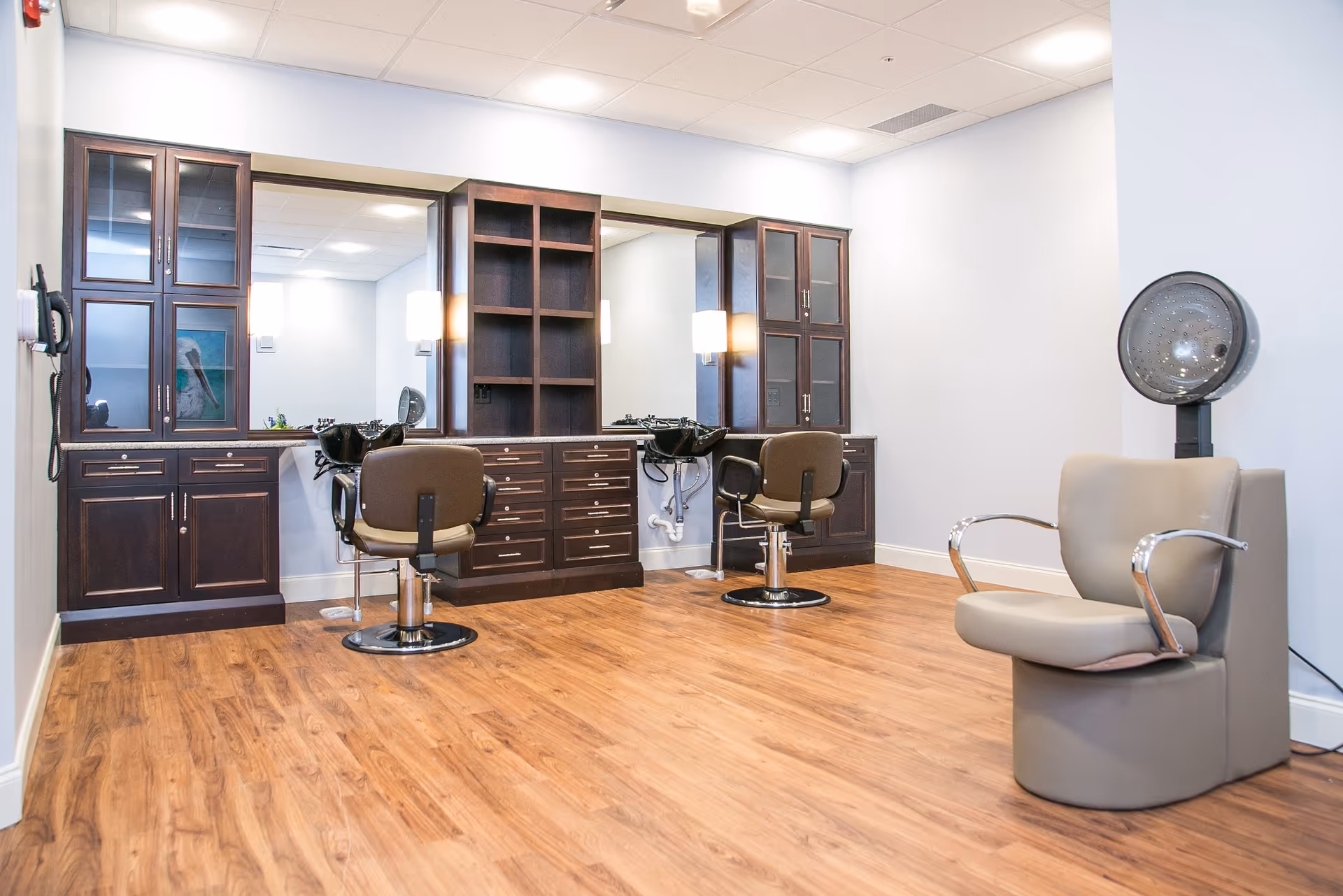 Modern salon area with styling chairs, mirrors, dark wood cabinetry and a hooded hair dryer on wood flooring.