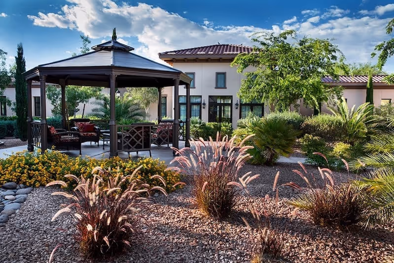 Outdoor garden area with a gazebo featuring seating and cushions, surrounded by various plants, shrubs, and trees. In the background, there is a building with multiple windows and a tiled roof under a partly cloudy blue sky.