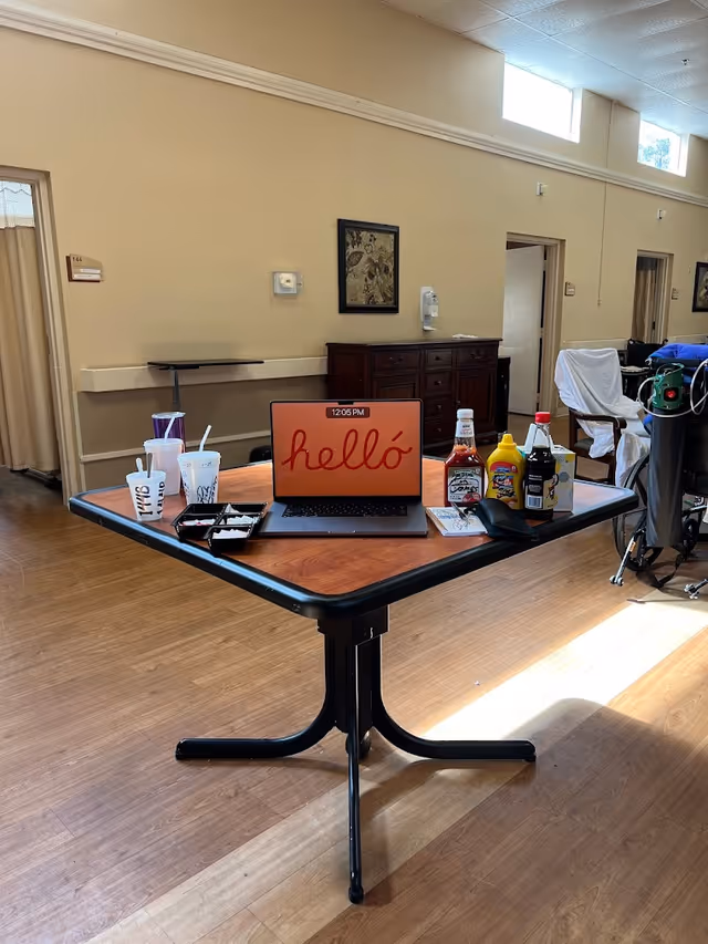 A communal indoor dining area with a table holding a laptop displaying "hello", drinks, condiments and takeout containers, with cabinets, chairs and medical equipment in the background.