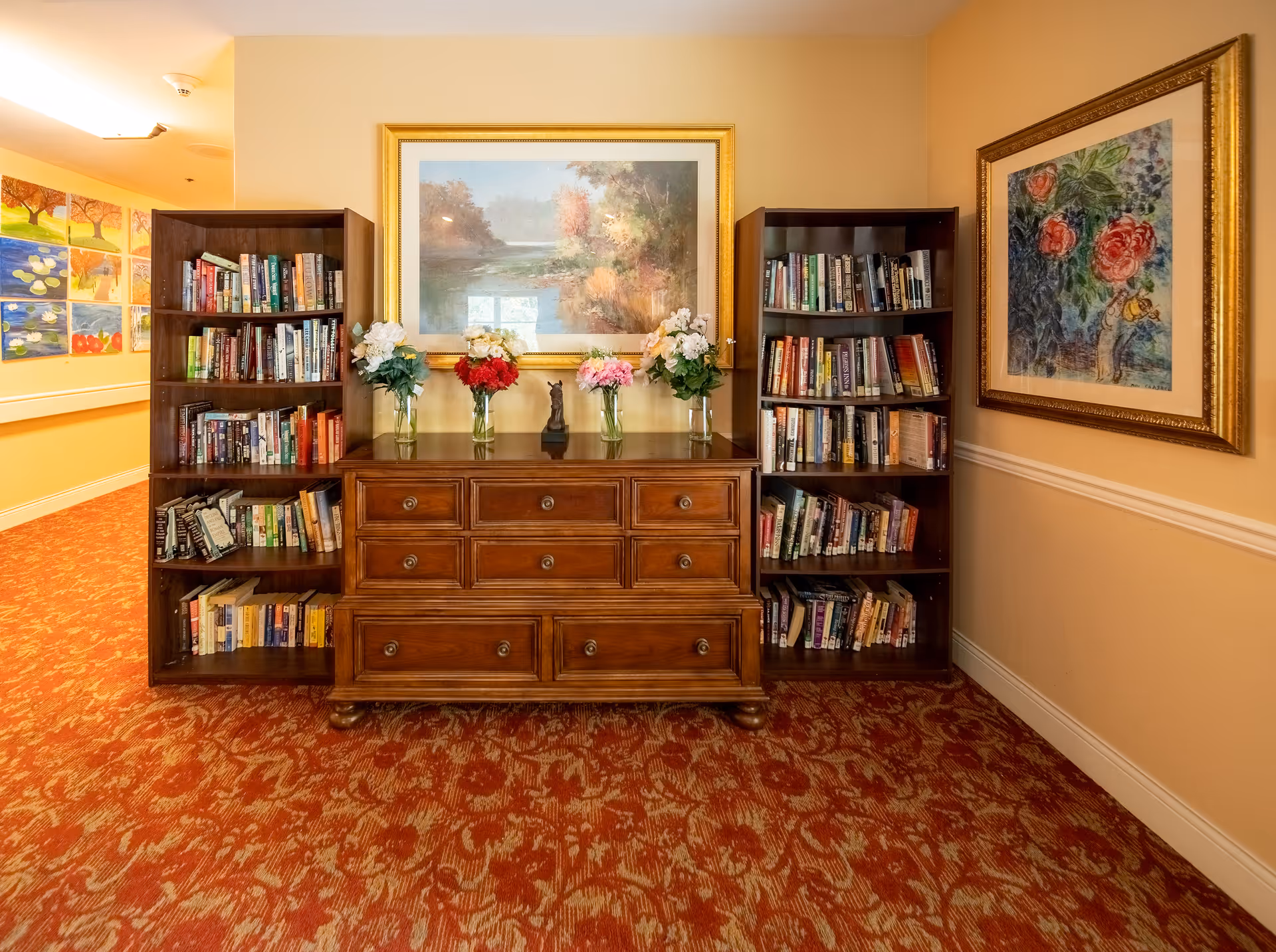 A cozy interior corner of a senior living facility featuring two wooden bookshelves filled with books on either side of a wooden chest of drawers. On top of the chest are five vases with colorful flowers and a small decorative statue. Above the chest hangs a large framed landscape painting, and to the right is another framed floral artwork. The floor is covered with a patterned red carpet, and the walls are painted a warm beige color.