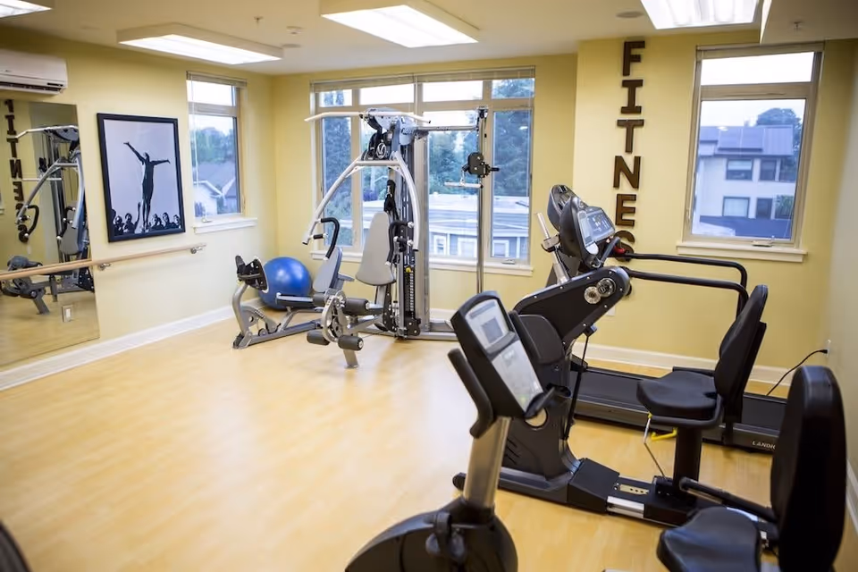 A bright fitness room with exercise equipment including a recumbent bike, a multi-function weight machine, a treadmill, and a blue exercise ball. The room has large windows letting in natural light, light wood flooring, and yellow walls. The word 'FITNESS' is displayed vertically on the wall near the windows, and a large mirror and framed artwork are visible on the opposite wall.