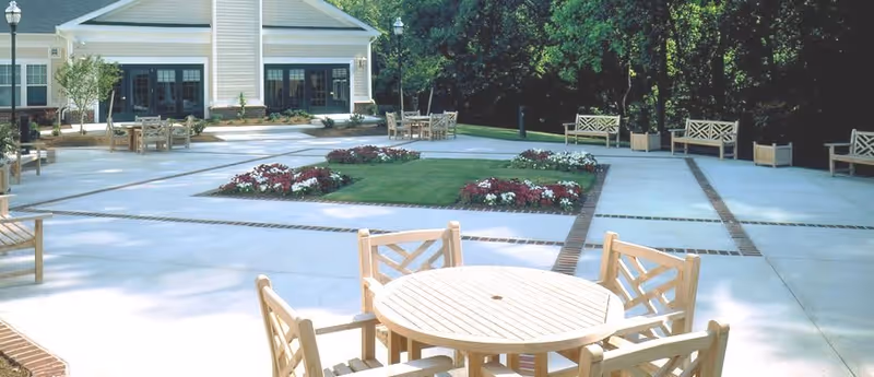 Outdoor patio area at St. Paul Senior Living with round wooden tables and chairs, benches along the perimeter, landscaped flower beds, and a building with large windows in the background.