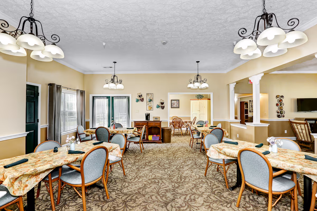 A bright dining room with multiple tables covered in floral tablecloths, each surrounded by chairs with light blue upholstery. The room has beige walls, patterned carpet, and several hanging light fixtures with white shades. There are windows with curtains and a set of double doors with glass panels. In the background, there is an adjacent room with more seating and a television mounted on the wall.