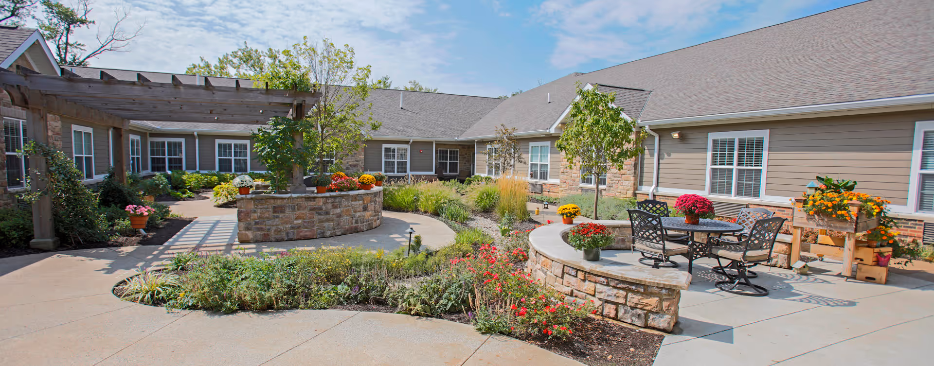 Sunny landscaped courtyard with stone planter walls, patio tables and chairs, and flowering plants in front of a single-story senior living building.