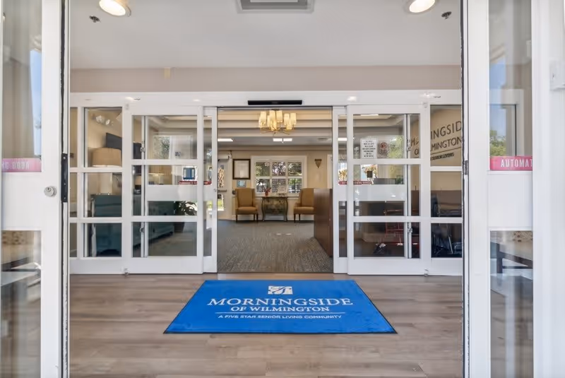 Entrance view into the Morningside of Wilmington senior living facility showing automatic glass doors opening into a lobby area with a blue floor mat displaying the facility's name. Inside, there are two armchairs and a small table with flowers, a chandelier overhead, and large windows letting in natural light.