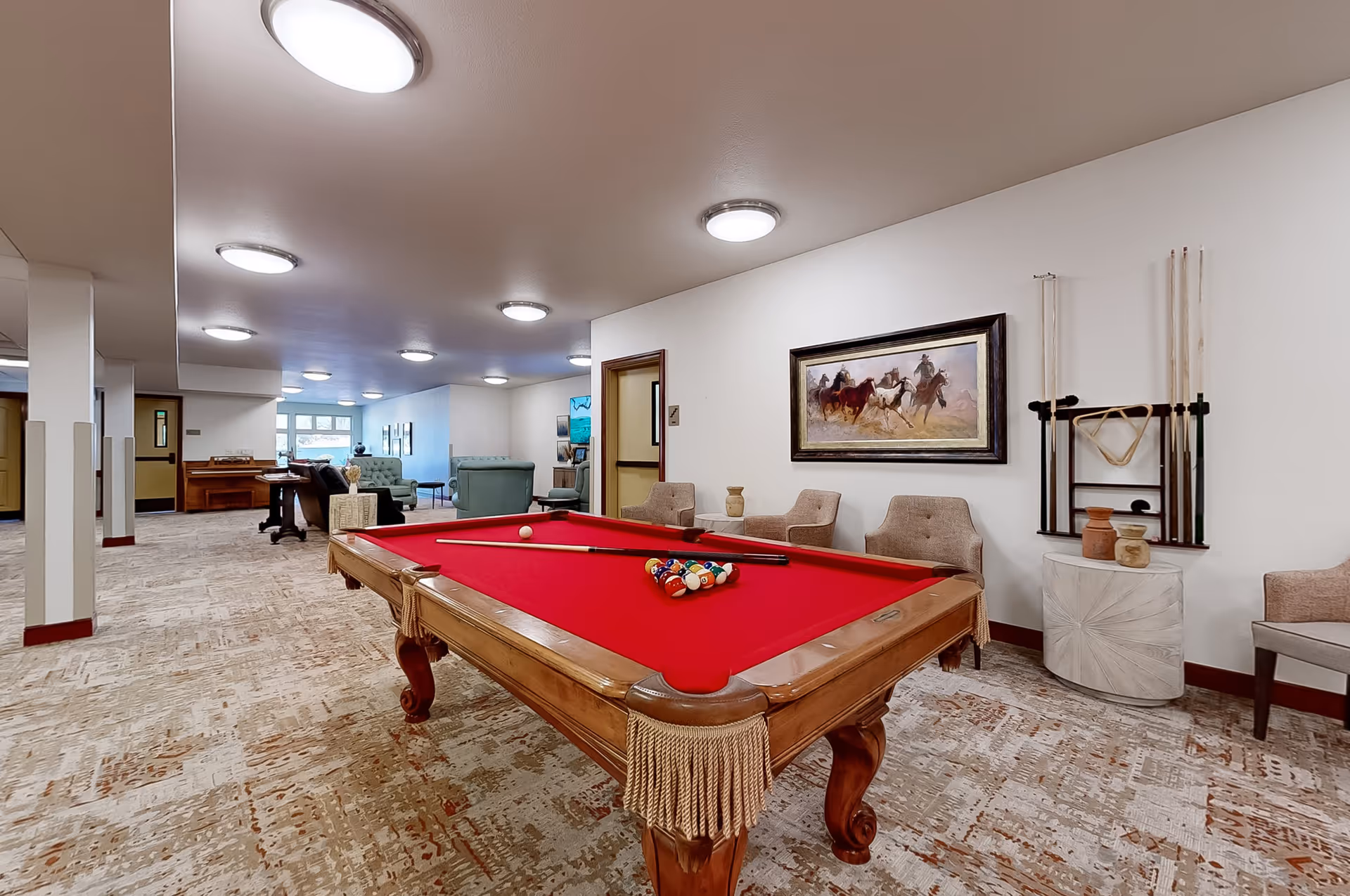 A spacious recreational room with a red felt pool table in the center, surrounded by beige chairs. On the wall behind the pool table, there is a painting of cowboys riding horses and a rack holding pool cues. In the background, there are several green upholstered chairs and a piano near a window letting in natural light. The room has a patterned carpet and multiple ceiling lights.