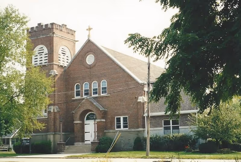 Exterior view of a brick church building with a cross on top of the front gable and a bell tower on the left side. The building is surrounded by trees and greenery.