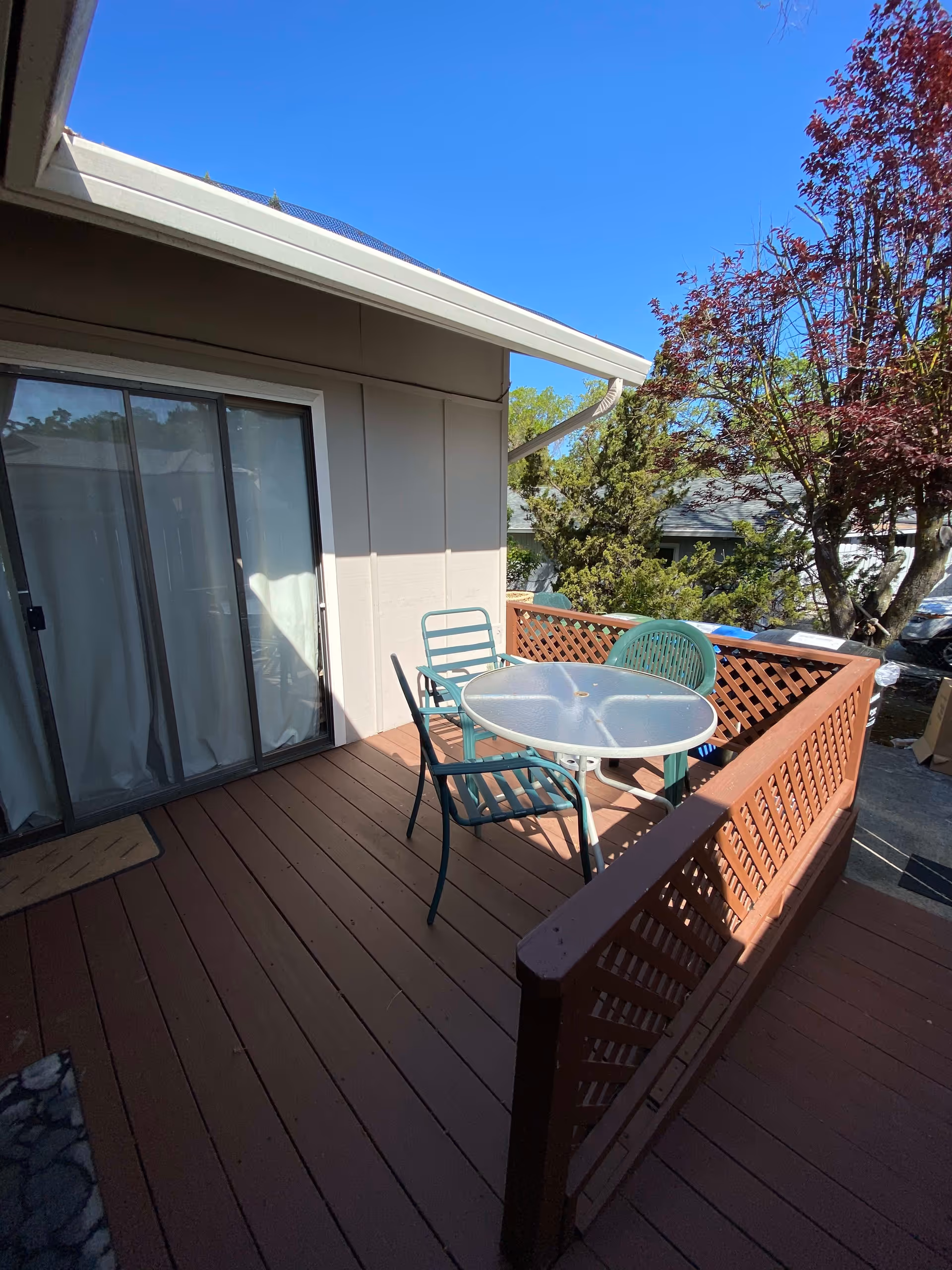 Outdoor wooden deck with a round glass table and three metal chairs, next to a sliding glass door. Trees and a clear blue sky are visible in the background.