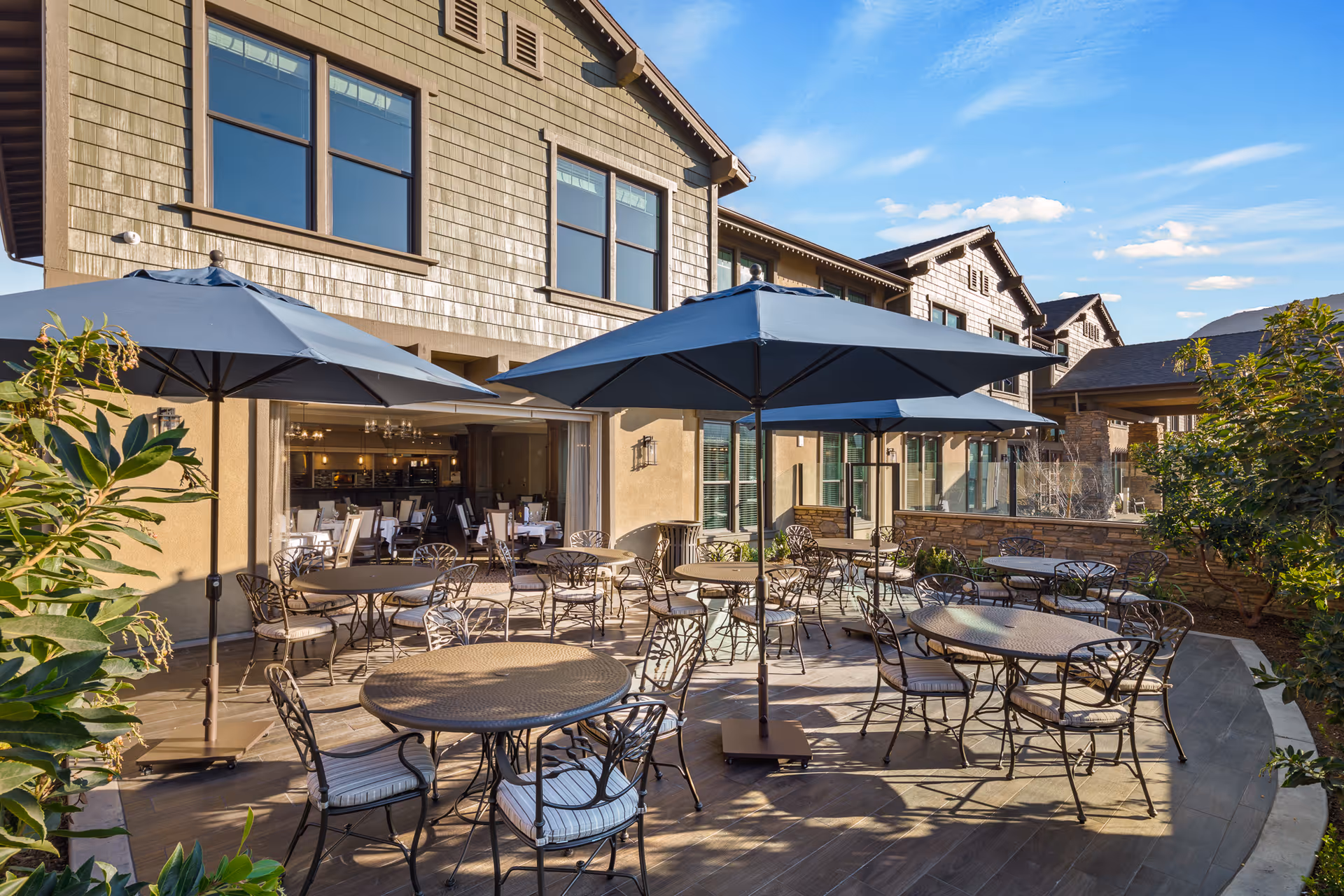 Outdoor patio area at Oakmont of Agoura Hills with several round metal tables and chairs, each shaded by large blue umbrellas. The patio is adjacent to a building with large windows and an open dining area visible inside. The sky is clear with a few clouds, and there are plants and greenery surrounding the patio.
