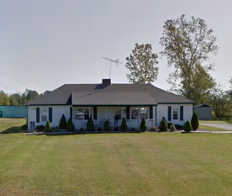 Single-story building with a covered front porch, manicured shrubs, and a large grassy yard.