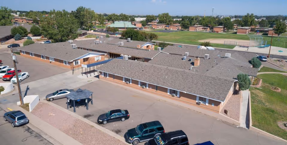 Aerial view of a single-story brick building complex with multiple connected sections and a gray shingled roof, surrounded by a parking lot with several parked cars. There is a small covered seating area in the parking lot, and green fields and trees are visible in the background under a clear blue sky.