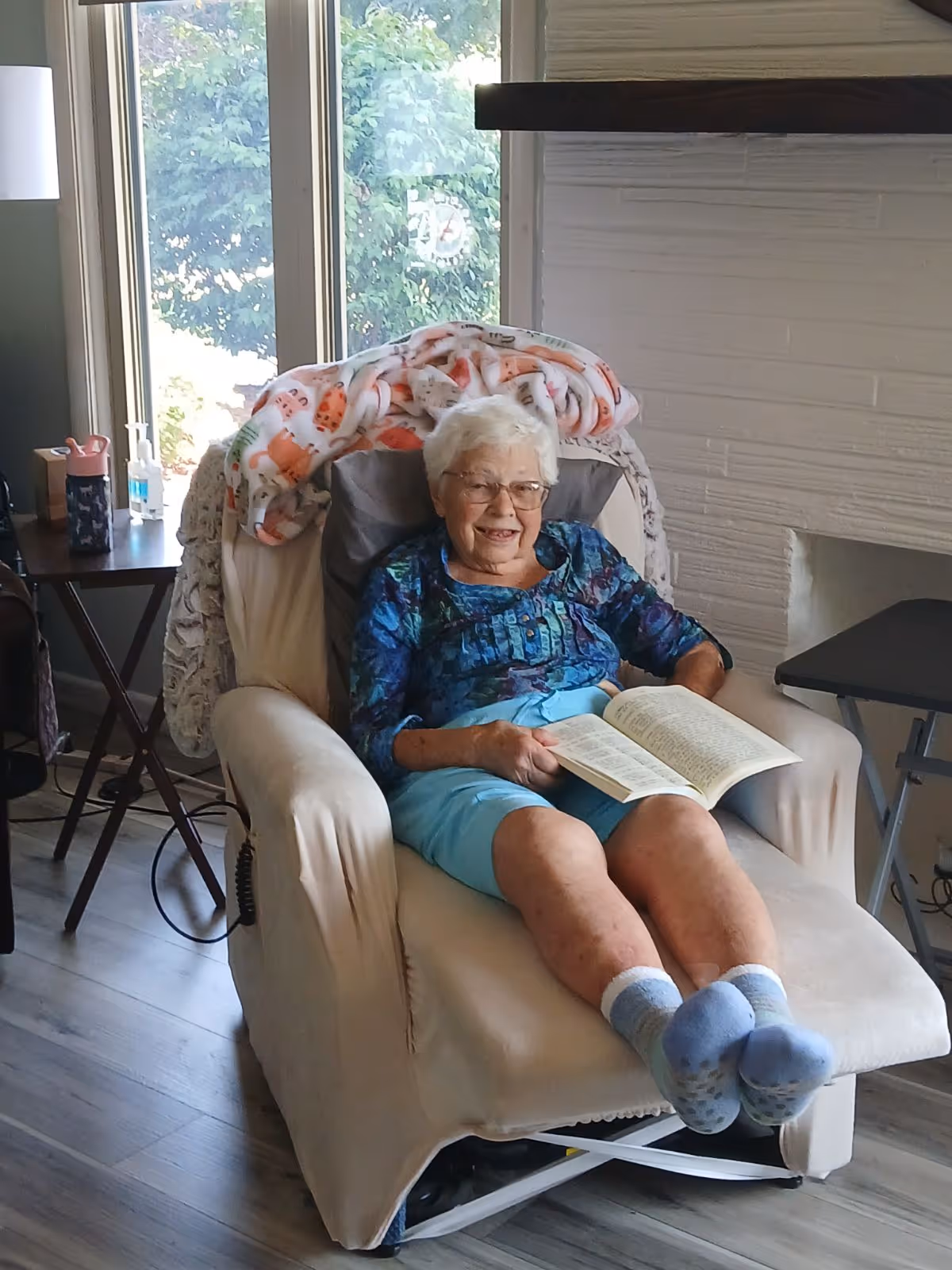 An elderly woman with white hair and glasses sitting comfortably in a beige recliner chair, reading a book. She is wearing a blue patterned top, light blue shorts, and blue socks. Behind her chair is a window with greenery outside, and a colorful blanket is draped over the back of the chair. There is a small table with a water bottle and hand sanitizer to the left, and a white brick fireplace to the right.