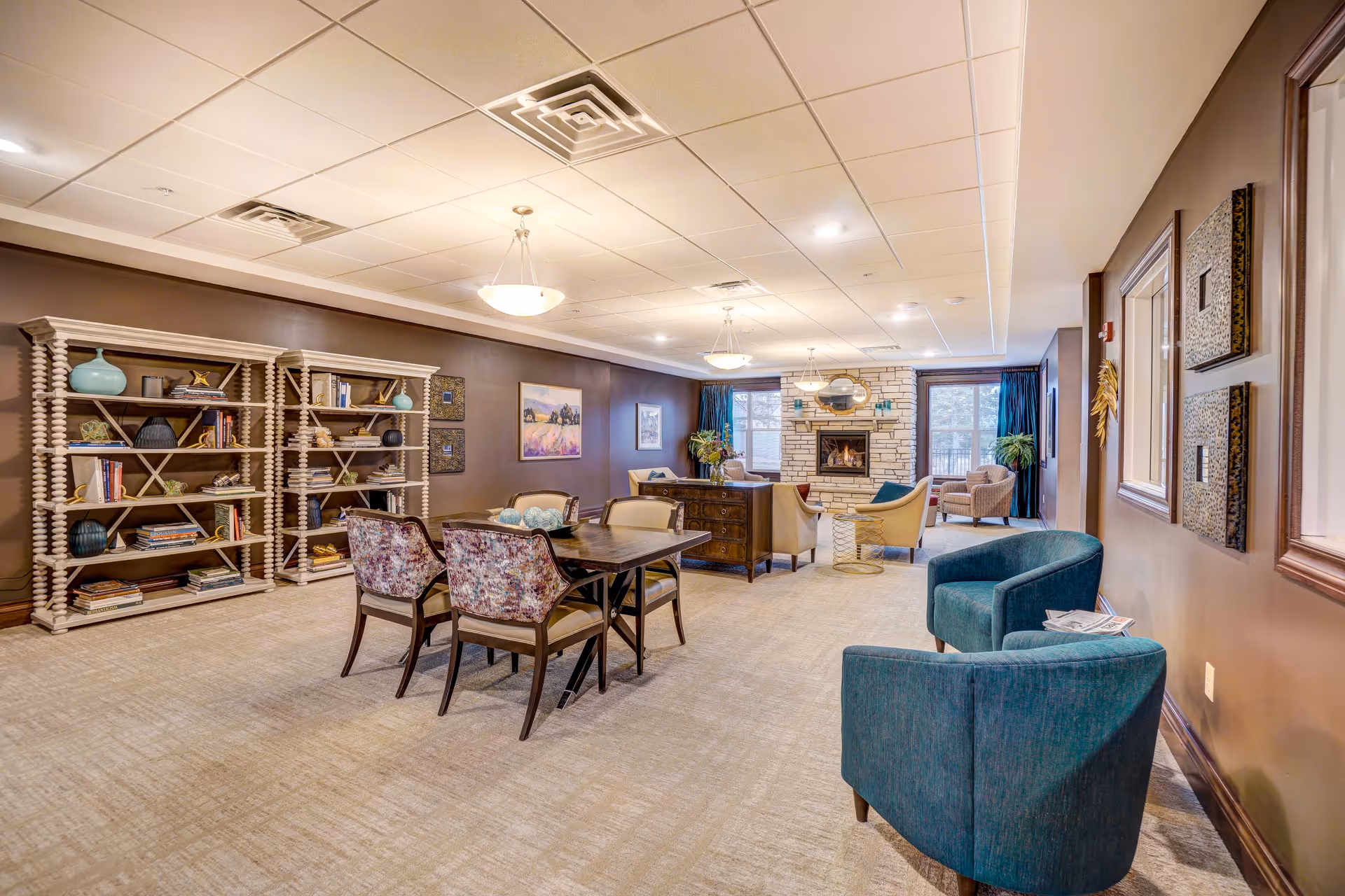 A spacious and well-lit living room area in a senior living facility with beige carpet, brown walls, and a white ceiling with recessed lighting. The room features a wooden table with four floral upholstered chairs, two teal armchairs near the wall, two large bookshelves filled with books and decorative items, and a cozy seating area with armchairs and a stone fireplace. Large windows with blue curtains allow natural light to enter the room.