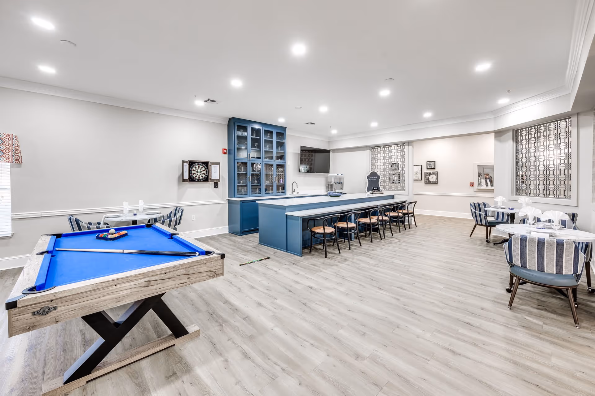 A spacious senior living common area featuring a blue pool table with cues and balls, a dartboard mounted on the wall, a long blue counter with several chairs, and multiple small round tables with striped chairs. The room has light wood flooring, white walls, recessed ceiling lights, and decorative window panels.