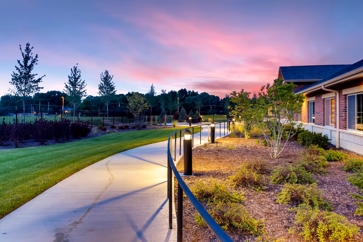 Curved concrete walkway with black railing and small lights along the side, leading past landscaped bushes and trees next to a brick and siding building under a colorful sunset sky.