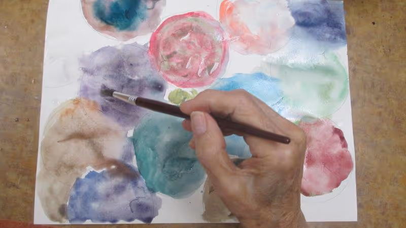 A close-up of an elderly person's hand holding a paintbrush, painting colorful circular shapes on a sheet of paper with watercolors.