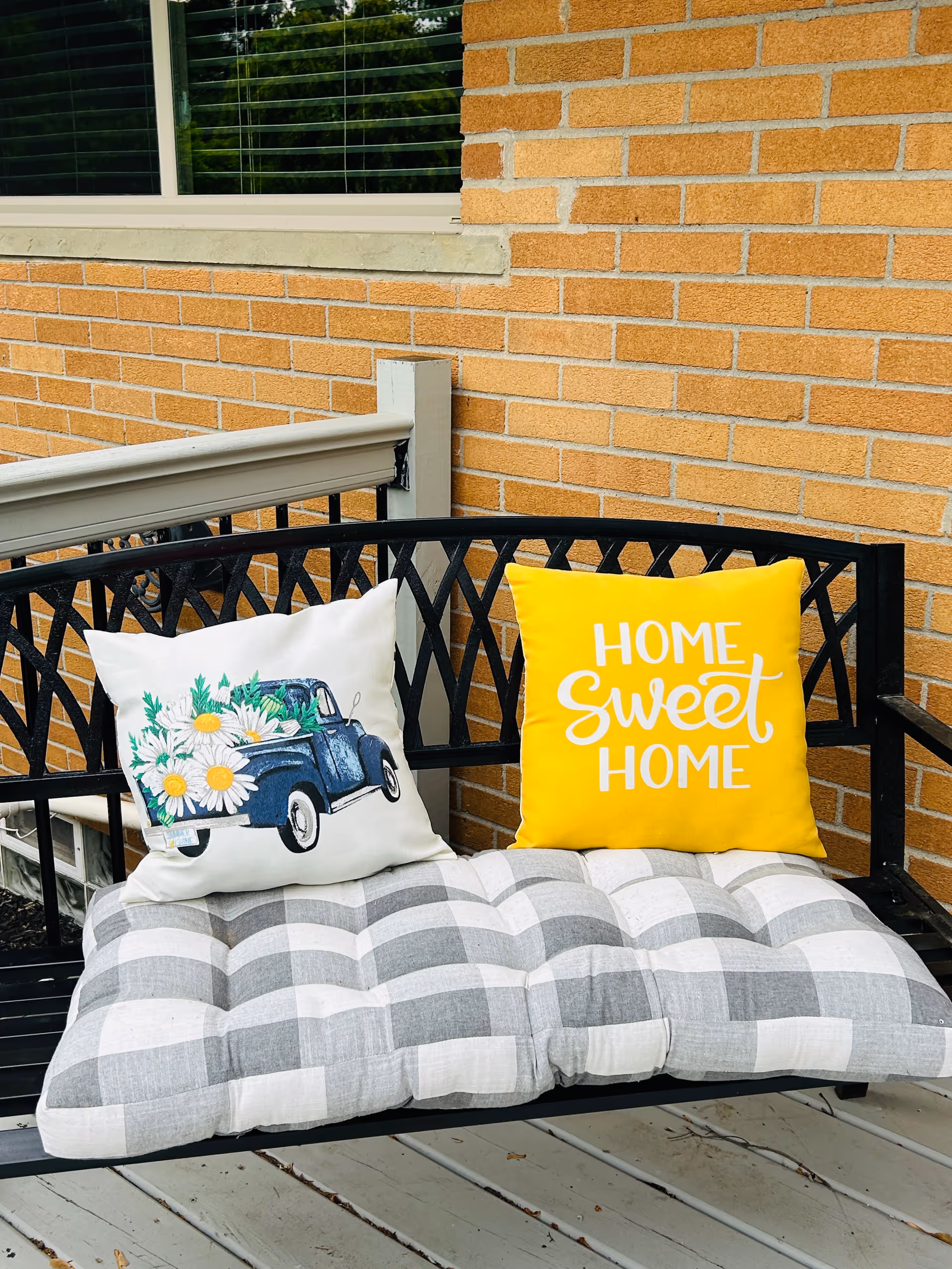Black metal bench with a gray and white checkered cushion and two decorative pillows, one with a blue truck carrying white daisies and the other yellow with the words 'HOME SWEET HOME', placed on a wooden porch in front of a brick wall and window with blinds.