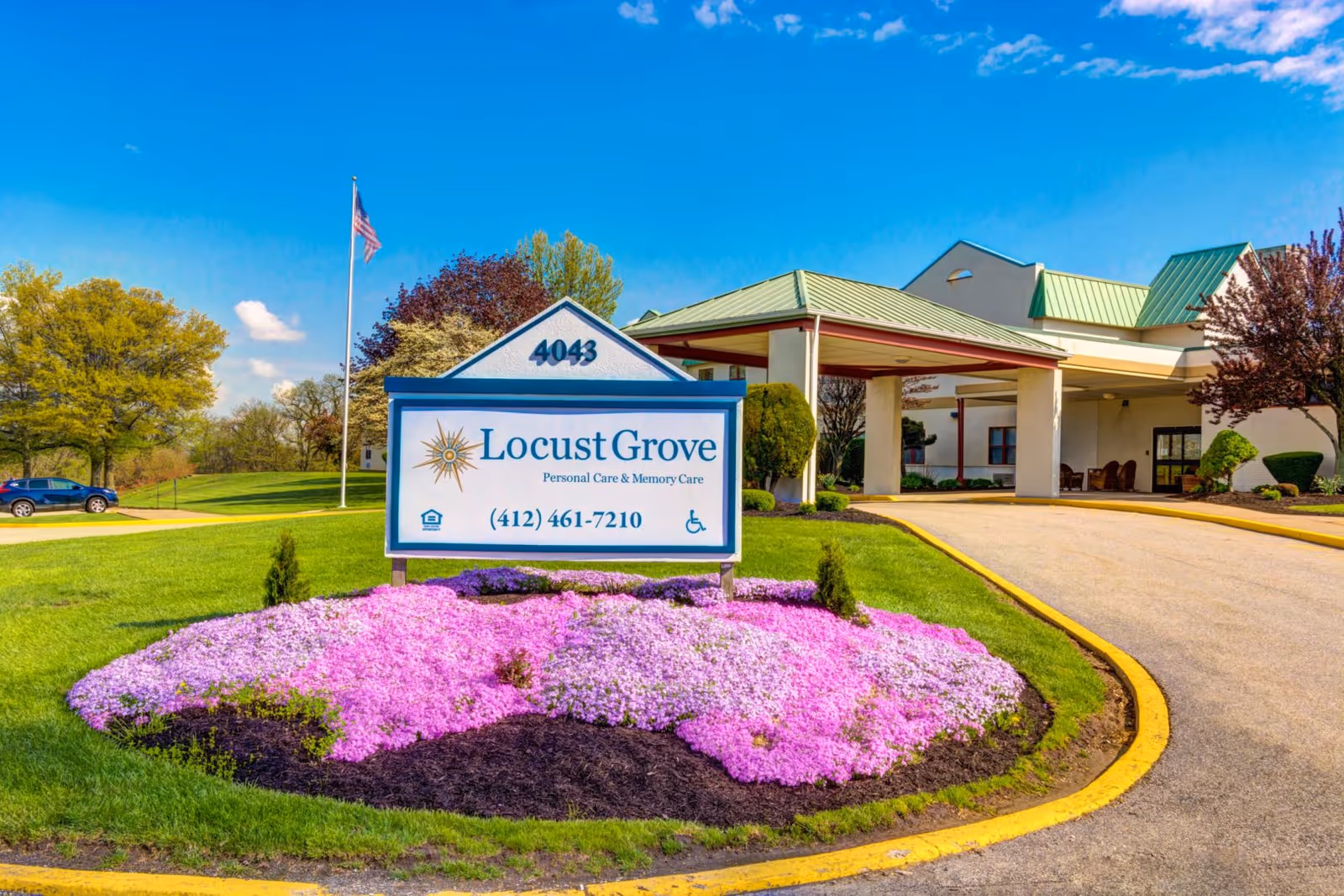 Entrance of Locust Grove Personal Care & Memory Care facility with a sign displaying the name, phone number, and address number 4043. The sign is surrounded by a flower bed with pink and purple flowers. The building has a green roof and a covered driveway. An American flag is visible in the background under a clear blue sky.