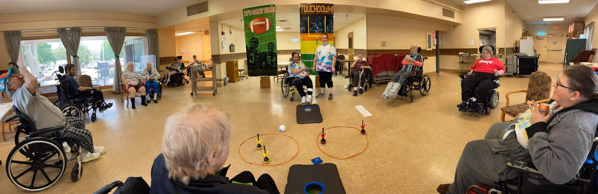 A group of elderly individuals, many in wheelchairs, seated in a large, well-lit room arranged in a circle. They appear to be participating in a recreational activity involving bean bags and rings on the floor. The room has large windows with curtains, beige walls, and banners with football themes hanging on the wall. A staff member stands among the group, facilitating the activity.
