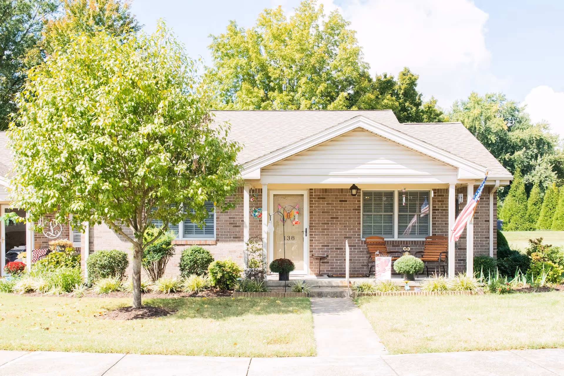 Front exterior view of a single-story brick home with a small porch, two wooden chairs, an American flag, and a tree in the front yard under a sunny sky.
