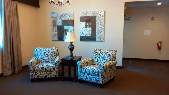Two patterned armchairs with a floral design are placed on either side of a small dark wooden table with a lamp on it. Above the chairs, there are two abstract paintings hanging on a beige wall. To the right, there is an open doorway leading to another area with a fire extinguisher mounted on the wall.