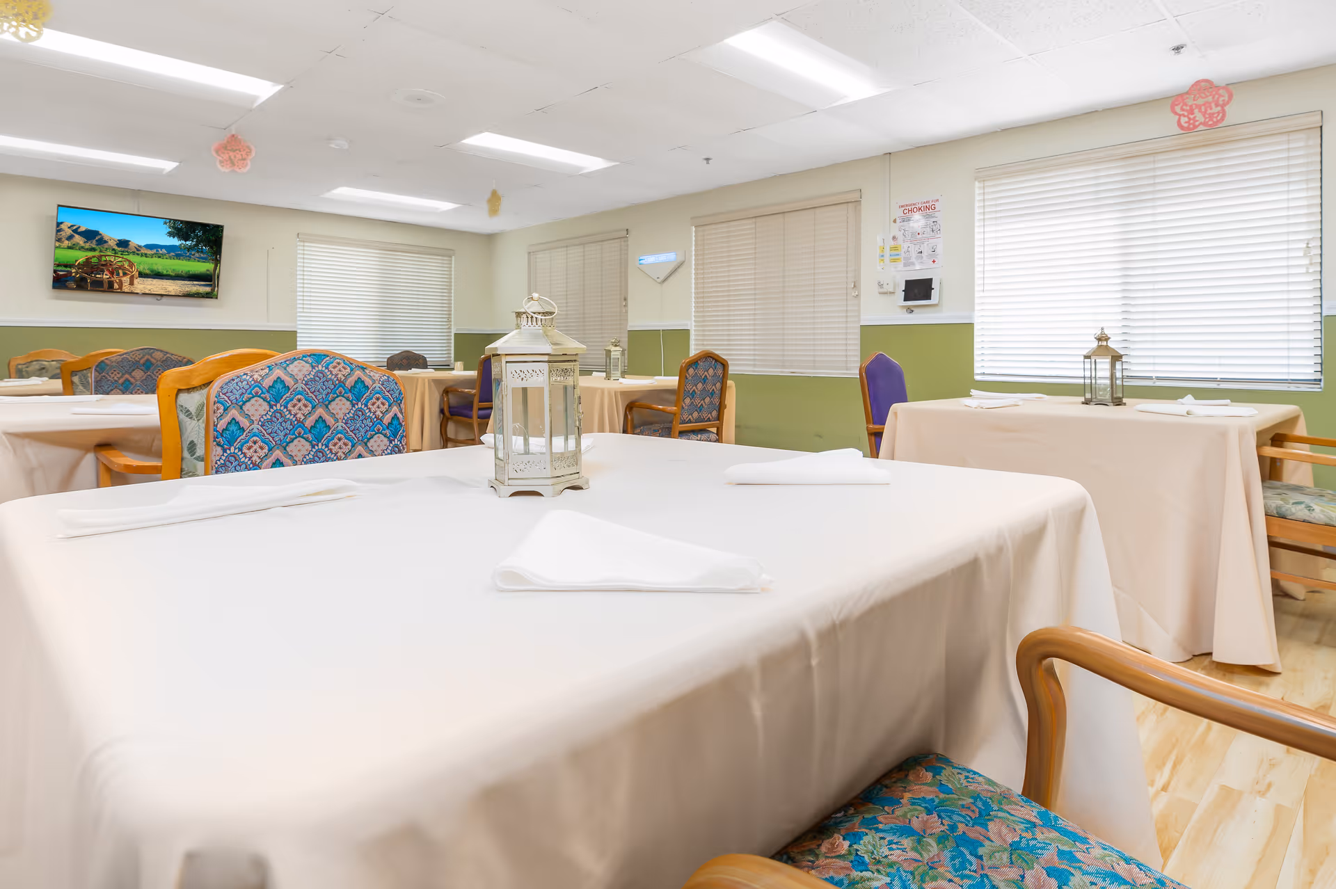 A dining room in a senior living facility with tables covered in white tablecloths and neatly folded napkins. The chairs have colorful patterned upholstery. Lantern-style centerpieces are placed on the tables. The room has large windows with blinds, fluorescent ceiling lights, and a wall-mounted TV displaying a scenic landscape.