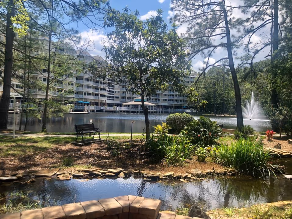 Landscaped pond and walking path with a bench and fountain in front of a multi-story Cypress Village building.