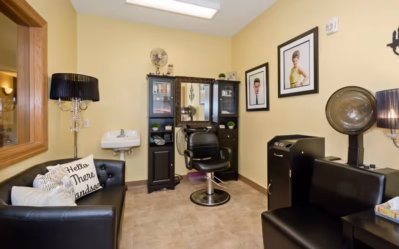 A small salon room with a black salon chair in front of a mirror and cabinet. There is a black leather couch with decorative pillows on the left, a black leather bench on the right, a hair dryer hood, a small sink on the wall, and two framed portraits hanging on the wall. The room has beige walls and tiled flooring.