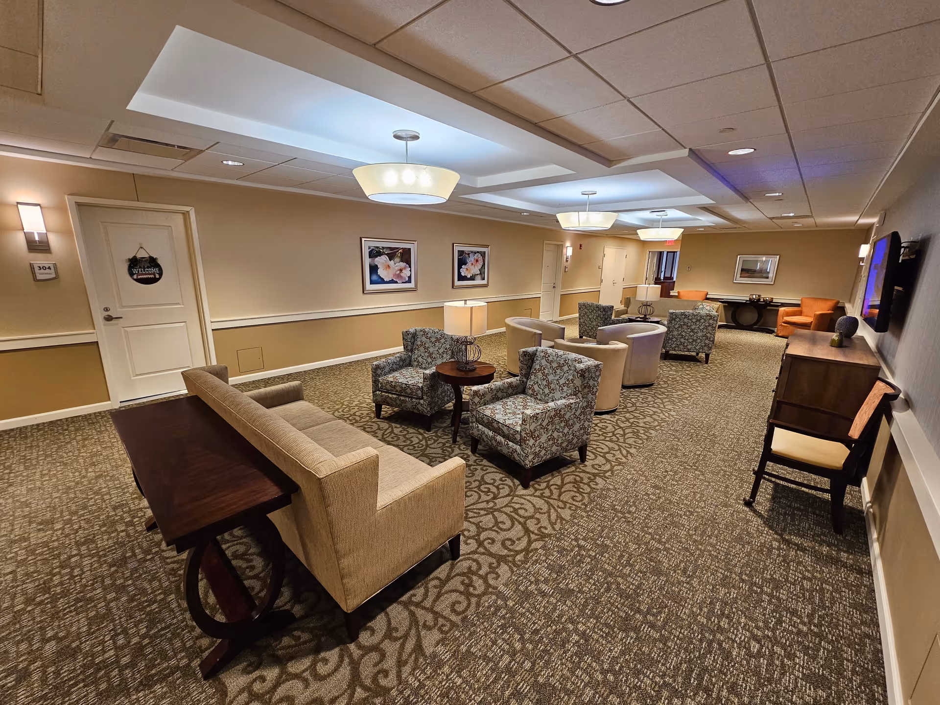 A spacious and well-lit common area in a senior living facility featuring multiple seating arrangements including beige sofas, patterned armchairs, and round tables with lamps. The walls are decorated with floral artwork, and the carpet has a patterned design. The ceiling has recessed lighting and modern light fixtures. Doors and a hallway are visible in the background.