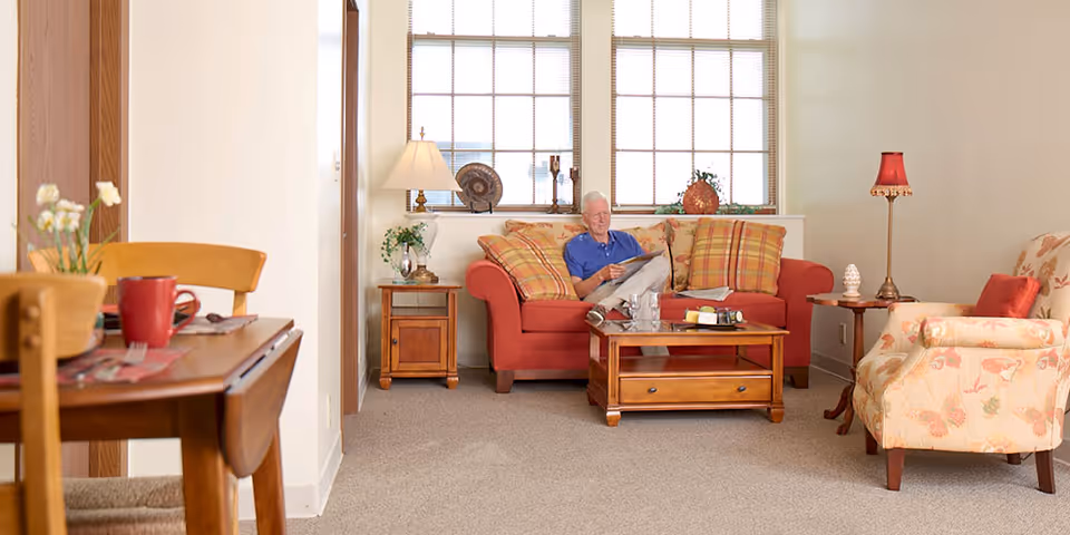 A senior man sitting on a red sofa in a cozy living room, reading a newspaper. The room features a wooden coffee table with books and a glass of water, a side table with a lamp and decorative items, a floral armchair with a red cushion, and a wooden dining table with a red mug and flowers in the foreground. Large windows with blinds let in natural light.