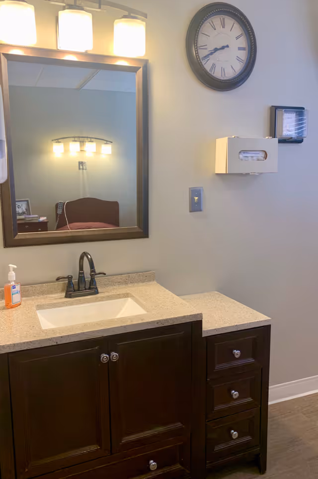 A bathroom vanity with a beige countertop, a white rectangular sink, and a dark wood cabinet with drawers. Above the sink is a large mirror with a dark frame, reflecting a bed and a wall light fixture. On the wall to the right of the mirror is a round clock with Roman numerals, a wall-mounted tissue dispenser, and a soap dispenser on the countertop.