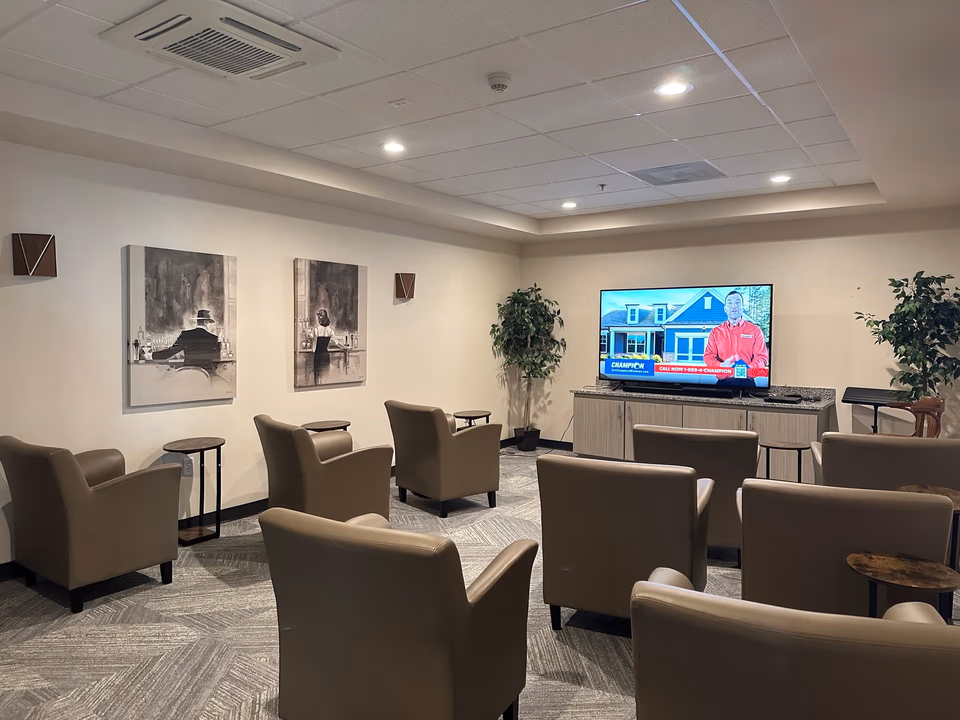 A cozy common area with several beige armchairs arranged facing a flat-screen TV mounted on a cabinet. The room has light-colored walls with two black and white paintings of people sitting at a bar. There are small side tables between some chairs and two potted plants near the TV. The ceiling has recessed lighting and an air vent.