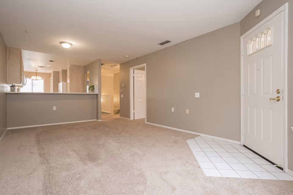 Empty interior room with beige carpet and gray walls, featuring a white front door with a small window, a tiled entryway, and a partial view of a kitchen area with a counter and cabinets in the background.