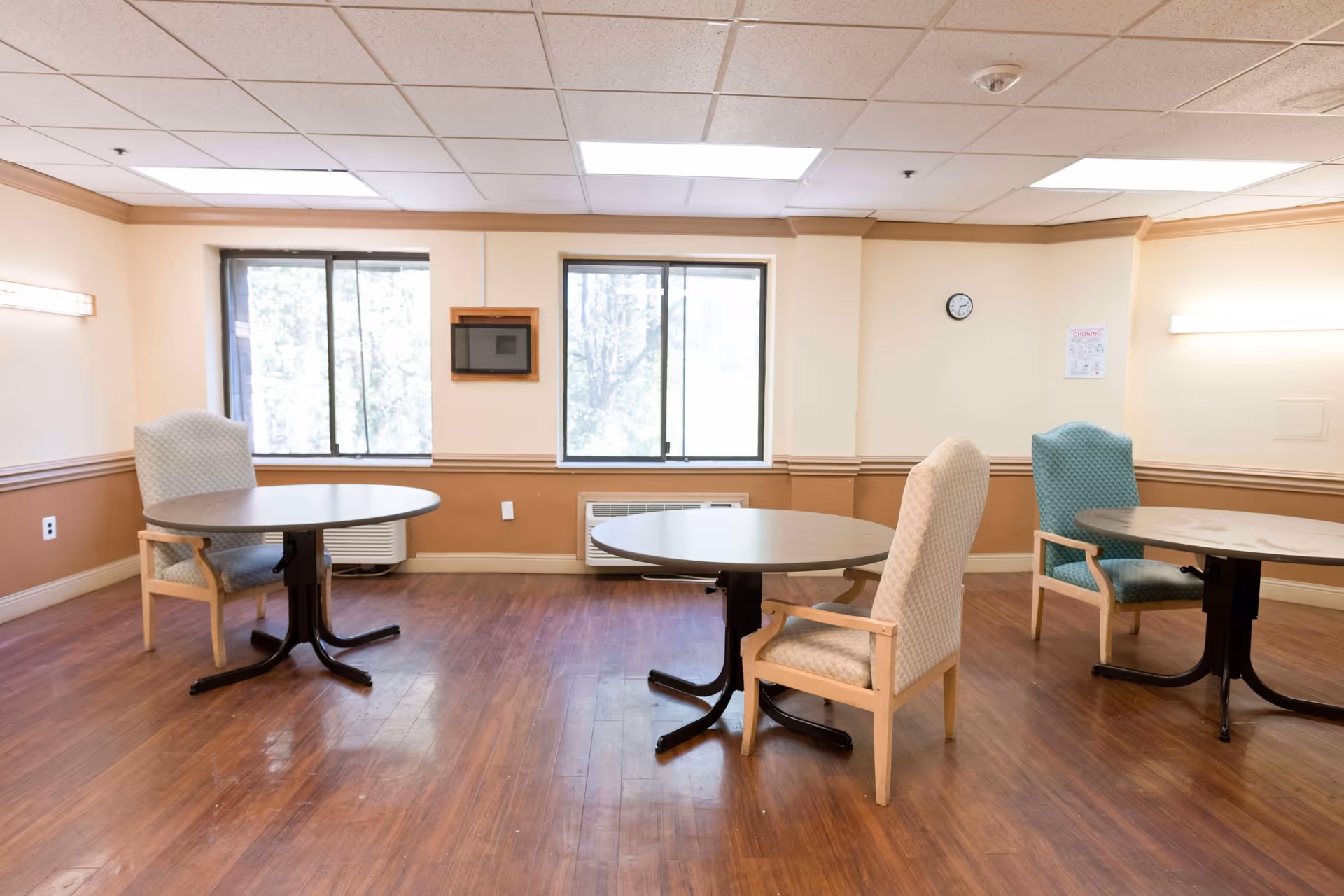 A room with three round tables, each accompanied by a cushioned armchair. The room has wood flooring, beige and brown walls, two large windows letting in natural light, a wall clock, and a small framed item on the wall.