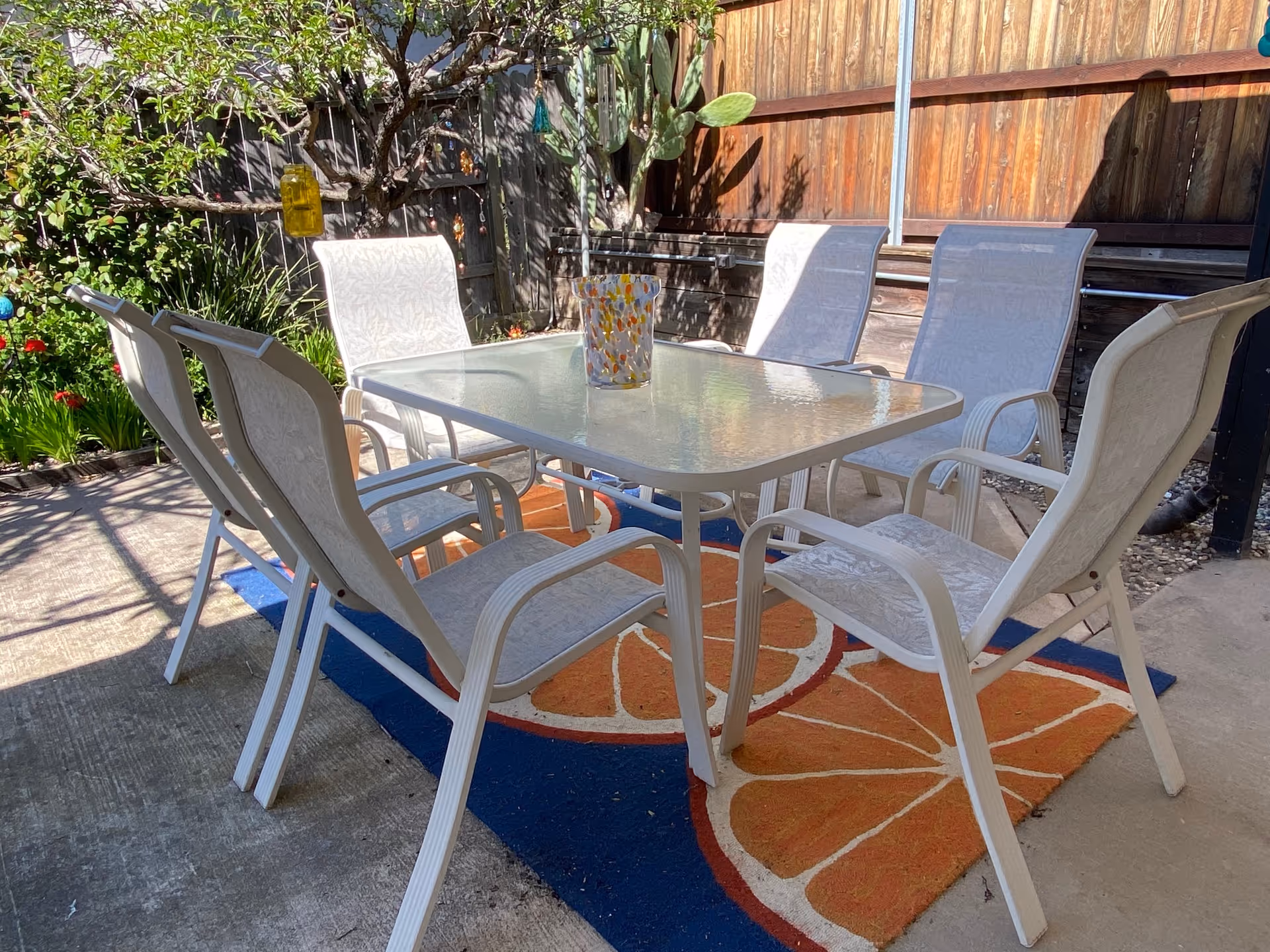 Outdoor patio area with a glass-top table surrounded by six white chairs on a colorful rug with orange slice designs. The area is enclosed by a wooden fence with plants and a tree in the background.