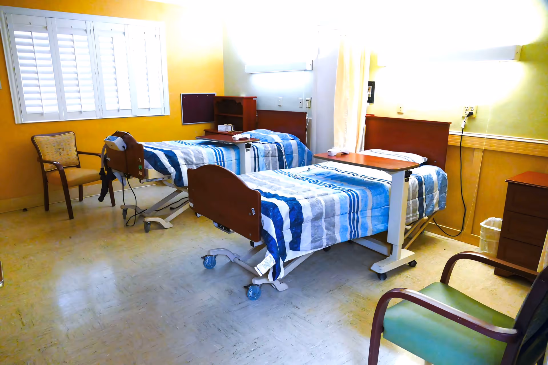 A hospital or rehabilitation room with two adjustable beds, each covered with blue and white striped bedding. The room has yellow walls, a window with white shutters, two chairs, a small wooden cabinet, and medical equipment attached to one of the beds.